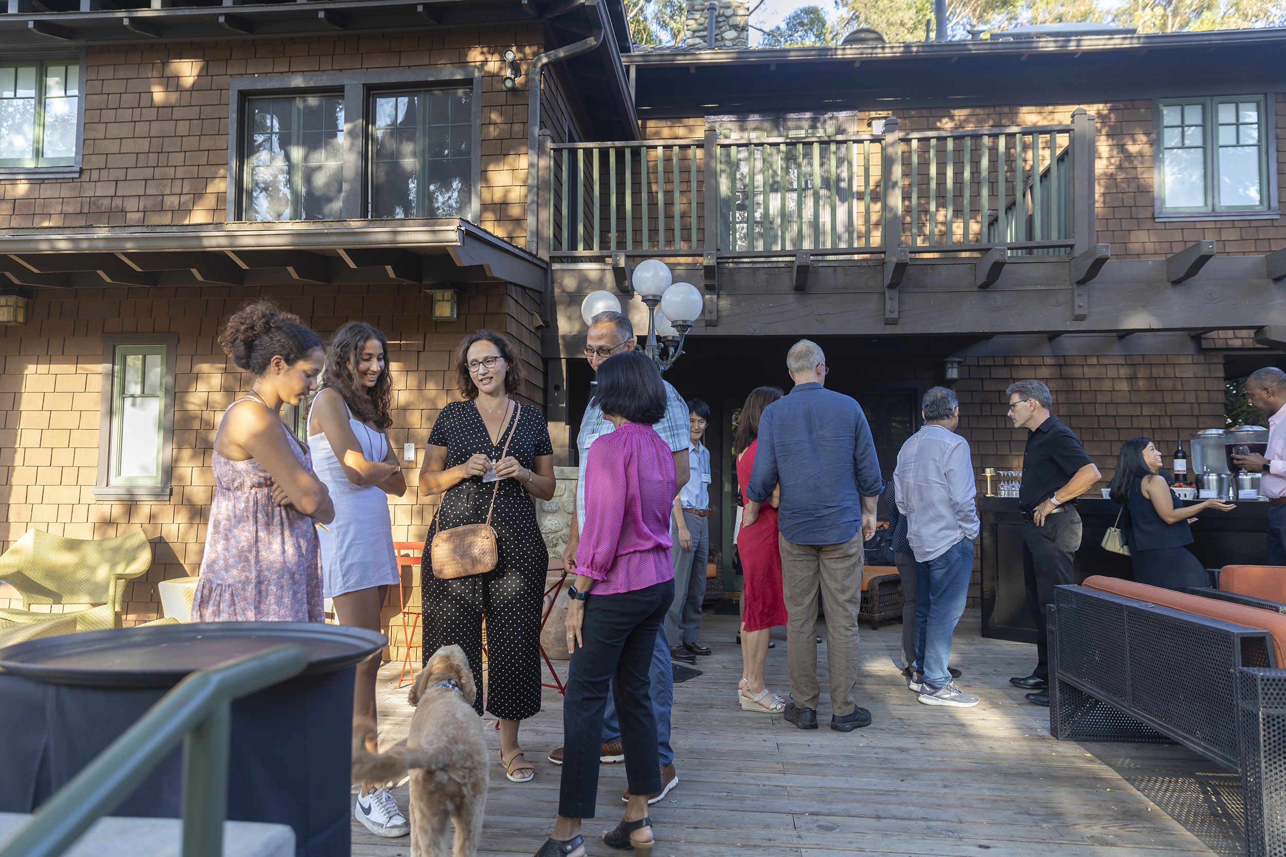 A circle of international students and their family mingle with staff at the President's sunny backyard. Joined by them is Ath Director Priya Junnar and her dog, Theo.