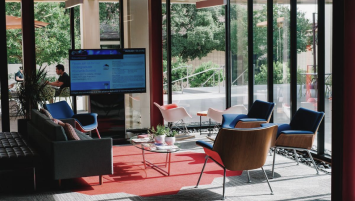 A media room with several chairs, a television, and a table, a view of green bushes behind through large windows