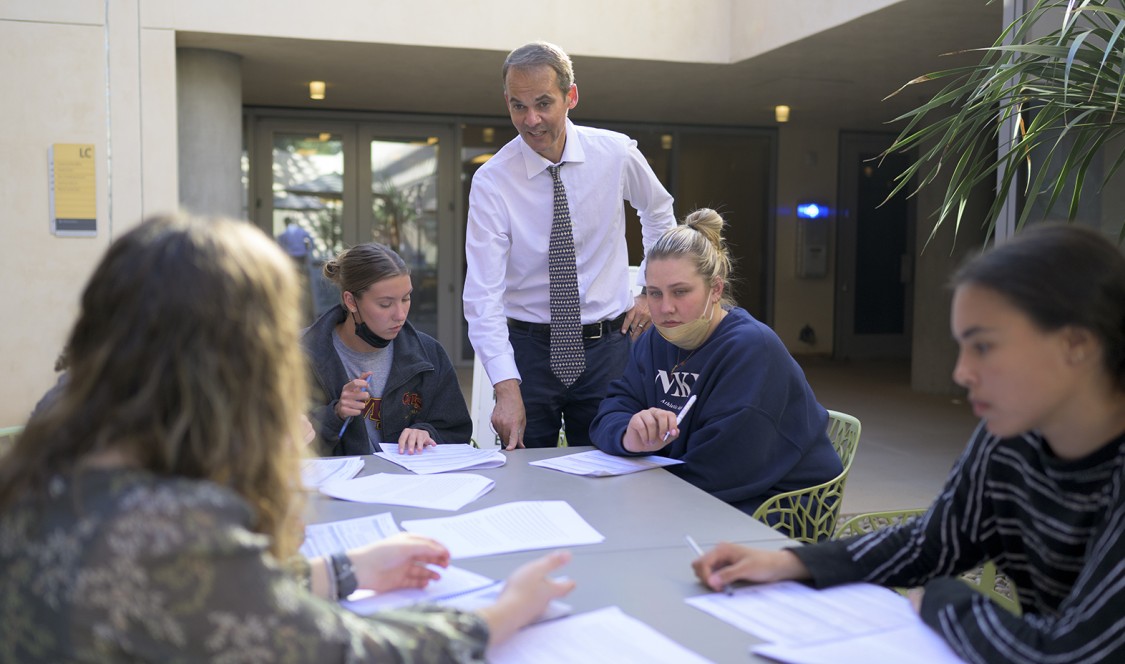 Candid photo of Daniel Krauss standing at an outdoor class setting with students seated around a table with papers.