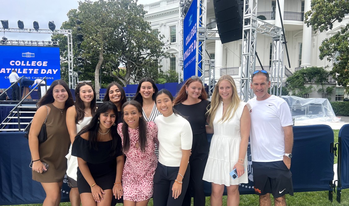 Group of female CMC tennis players and their coach stand and squat in front of a blue stage set up next to the White House for the event.