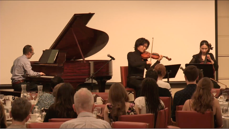 Photo of a man playing a grand piano, with two violinists playing next to him, on stage at the Athenaeum