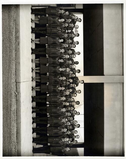 Group of men and women in military uniforms in front of gray structure. 