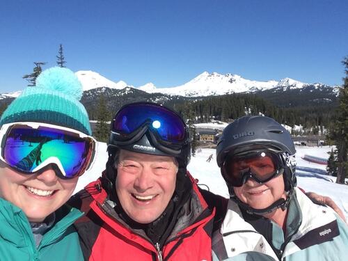 Man in red snow outfit with two women in blue and white snow outfits and snow glasses standing on snow in mountains.