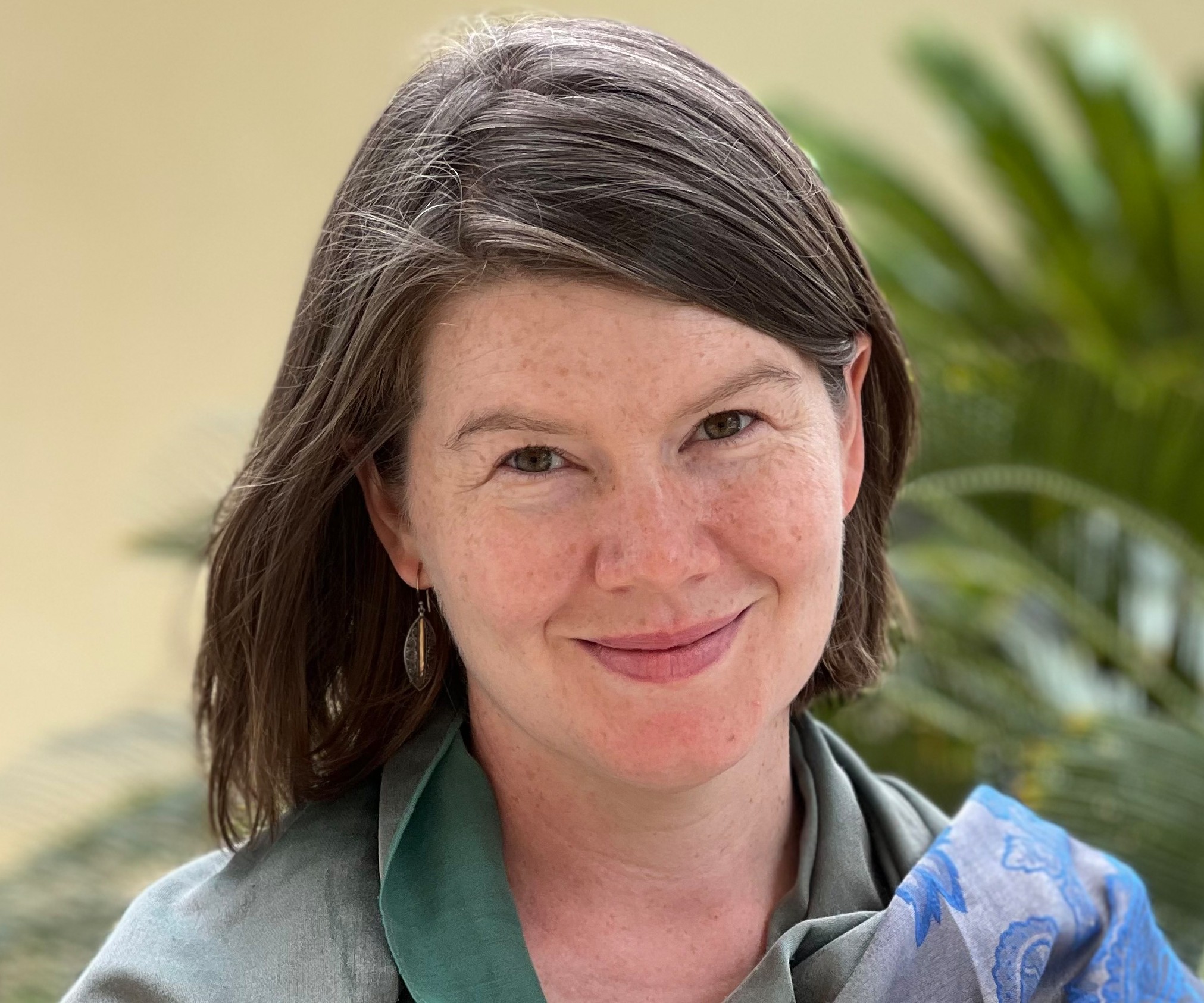 Photo of Gabby Salazar, a light-skinned woman with freckles and dark hair, wearing a series of blue and gold patterned scarves over a green top.