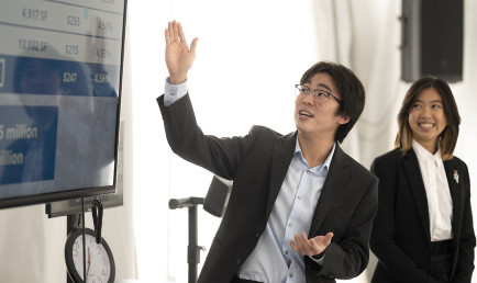 Two students present their pitches using a screen monitor as an analog clock in the back times them.