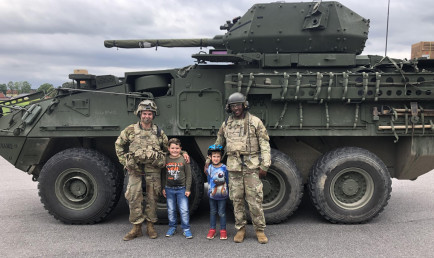 Ash and his friend stand with two children in front of a tank.