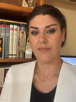 Woman with short brown hair wearing white blazer and black top in front of library.